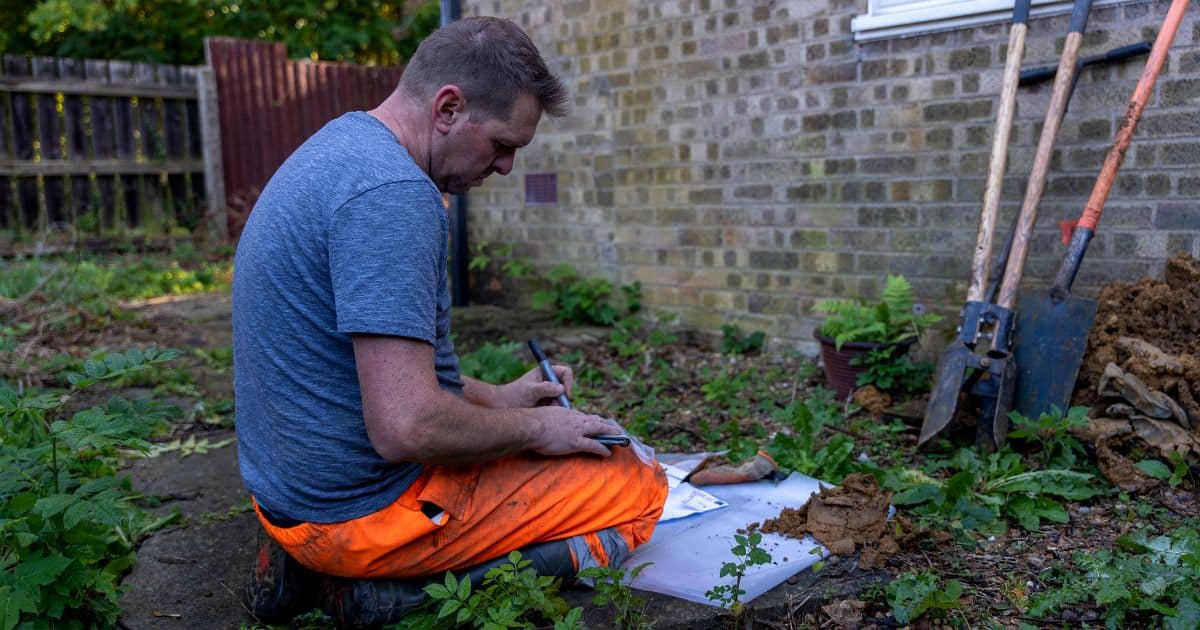 Structural engineer recording soil samples beside a trial pit dug next to a property's foundations, with spade and probe rods against the wall